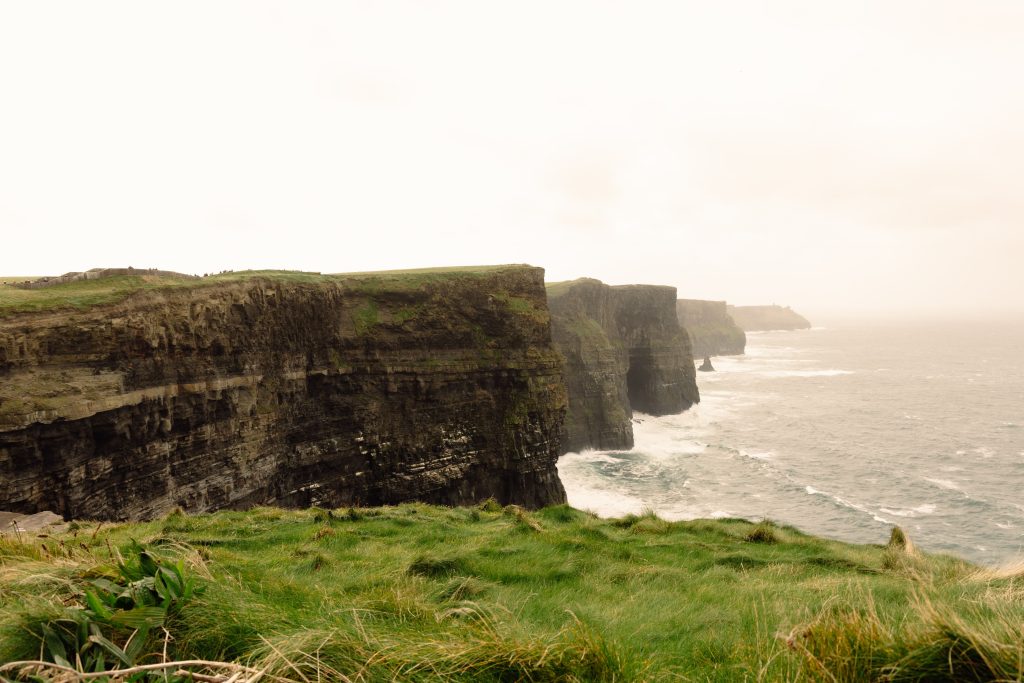 Dark layered cliffs dropping into the Atlantic, waves breaking below, green grass in the foreground and visitors on a clifftop path in the distance.